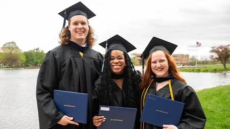 Three college students in graduation regalia smiling by lake